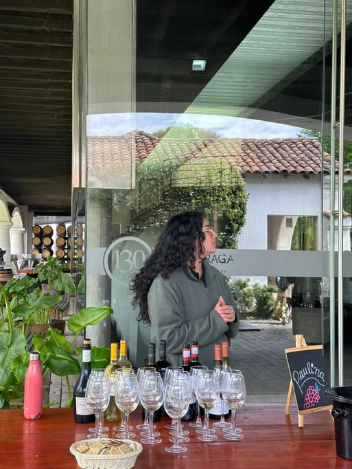Mulher com cabelos cacheados escuros usando jaqueta cinza, de cabelos ao pescoço, de perfil, dentro de uma loja de vinhos, ao lado de uma mesa com várias taças de vinho, garrafas de vinho, uma cesta com pão e uma carta de vinhos.