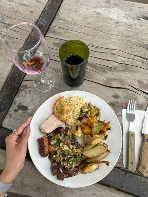 Prato com purê de batata, carne assada, picadinho com salsa, vegetais cozidos e fatias de pão, acompanhado de uma taça de vinho tinto, uma taça vazia e uma bebida escura, sobre uma mesa de madeira ao ar livre.