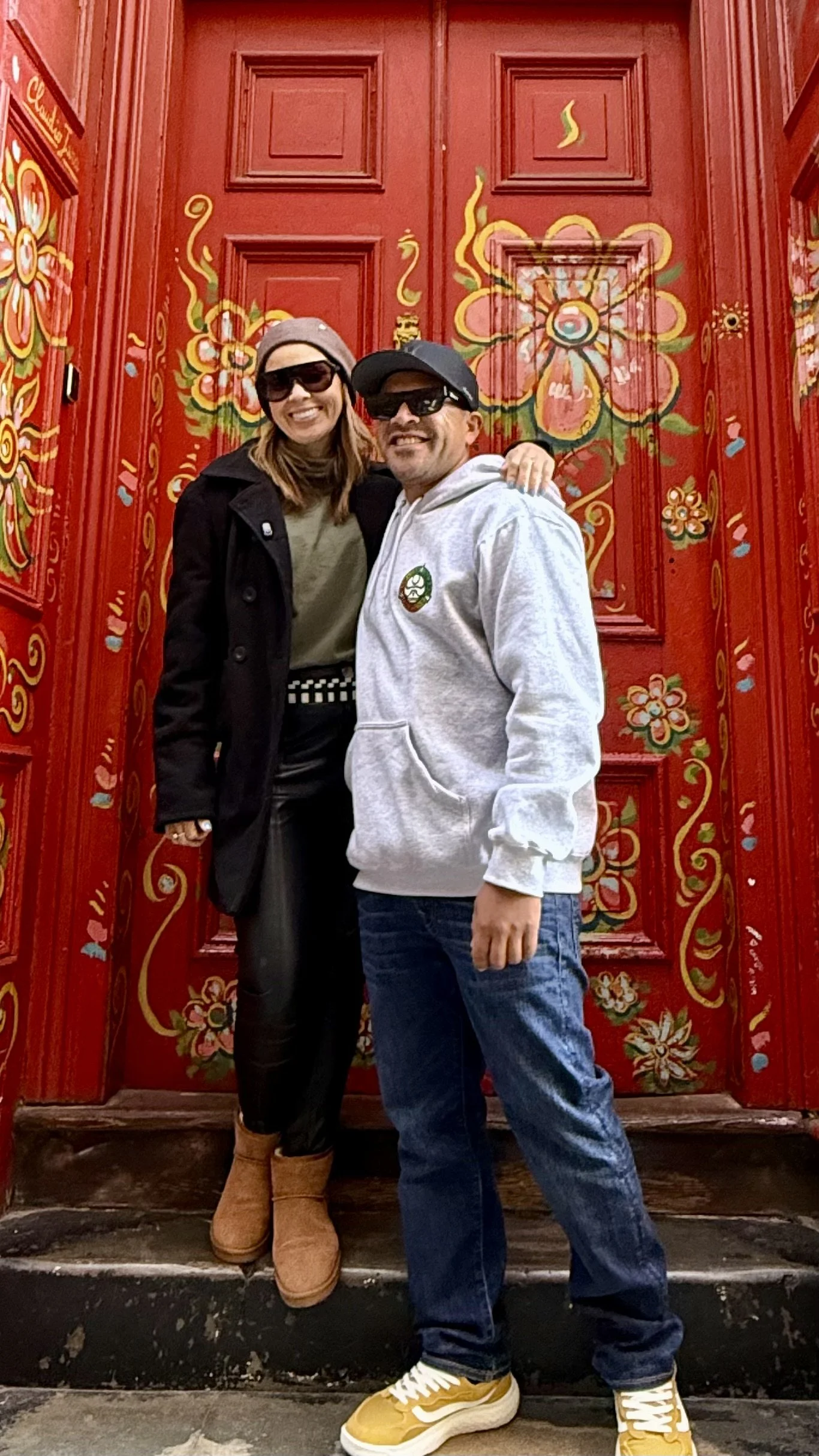 Pessoa vestida com casaco listrado preto e branco, calças pretas, óculos escuros e chapéu, de pé na porta vermelha decorada com desenhos florais, com graffiti colorido nas paredes ao redor.