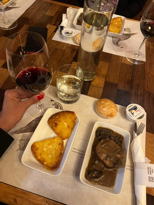 Prato com dois pedaços de pão de queijo, um pedaço de pão comum, carne com molho e uma taça de vinho tinto, uma garrafa de água e um copo de água sobre a mesa de madeira