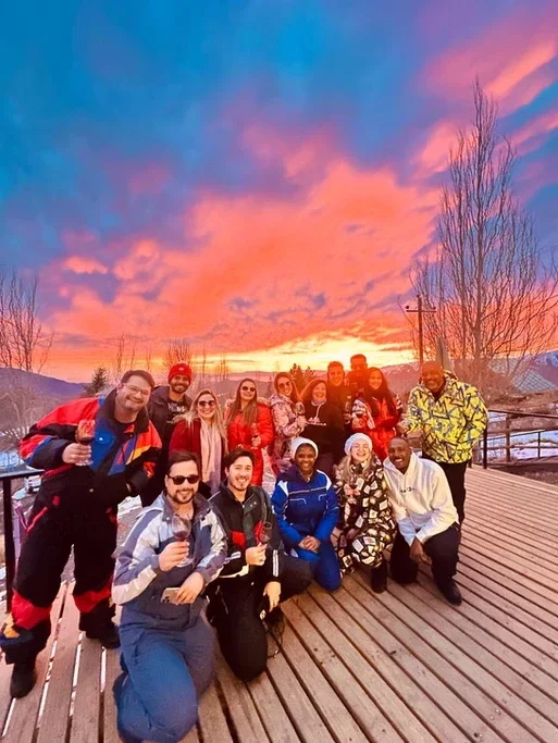 Grupo de pessoas sorrindo ao pôr do sol em uma ponte de madeira ao ar livre.