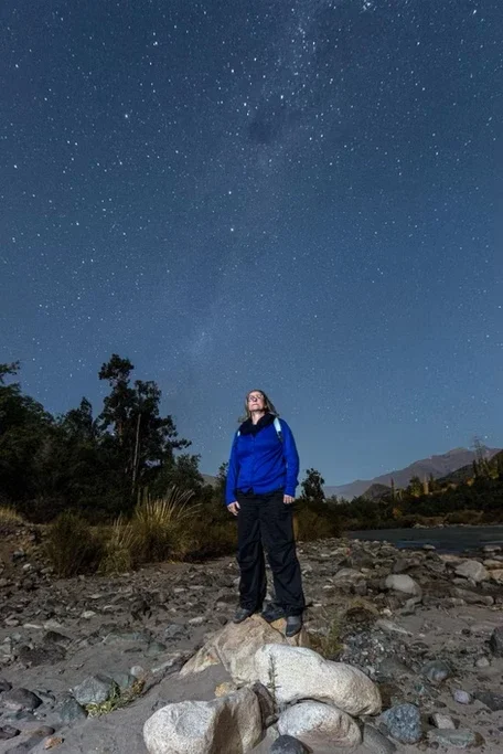 Pessoa observando o céu estrelado em uma paisagem natural com árvores, montanhas e rio.