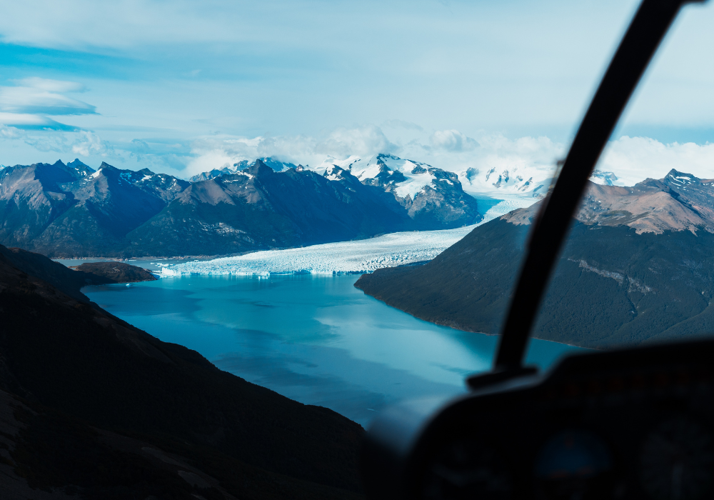 Vista de uma geleira e montanhas ao fundo, vista de um helicoptero, com o cockpit parcialmente visível.