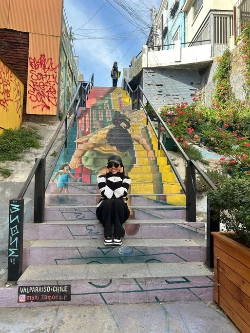 Pessoa sentada na escada decorada com arte colorida de rua, com uma criança pintada a poucos passos, e uma mulher ao topo da escada, enxerga a rua. Flores e edifícios fazem parte do cenário.