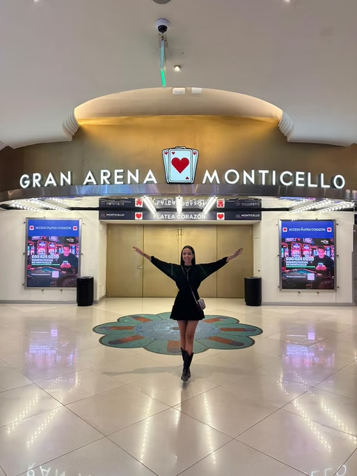 Pessoa posando de braços abertos na entrada do Gran Arena Monticello, com duas telas ao fundo exibindo imagens e o nome do local acima da porta.