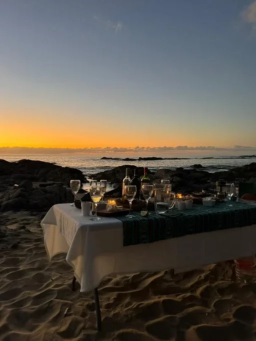 Mesa de jantar ao ar livre na praia ao pôr do sol, com copos, garrafas e velas
