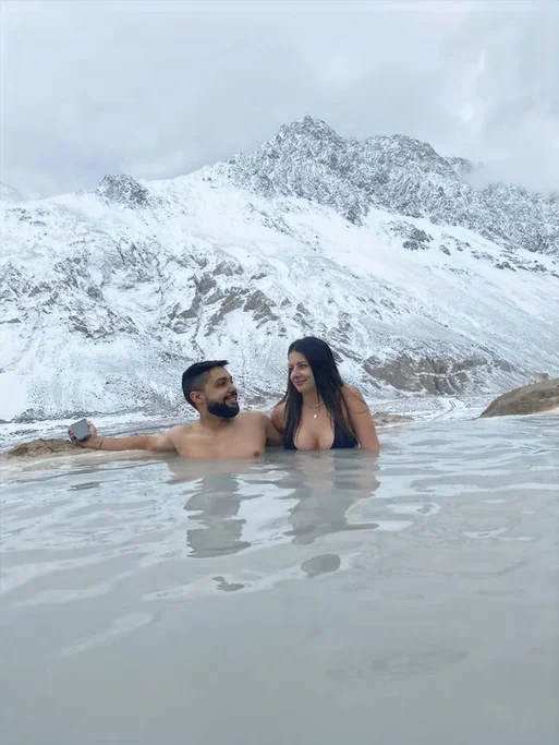 Casal tomando banho termal em uma piscina natural com montanhas nevadas ao fundo.