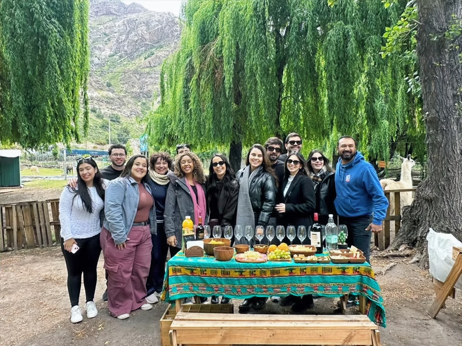 Grupo de pessoas sorrindo ao redor de uma mesa de piquenique com comidas e bebidas, cercados por árvores verdes e um cenário de montanhas ao fundo.