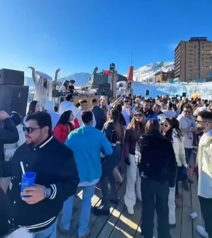 Pessoas em um evento ao ar livre, com decoração temática de inverno e montar de neve artificial, ao fundo montanhas de neve e edifícios em um dia ensolarado.