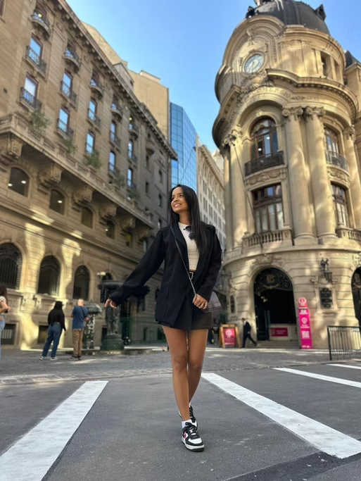 Mulher sorridente caminhando na rua, vestindo saia curta, jaqueta escura e tênis, com edifícios históricos ao fundo em uma cidade.