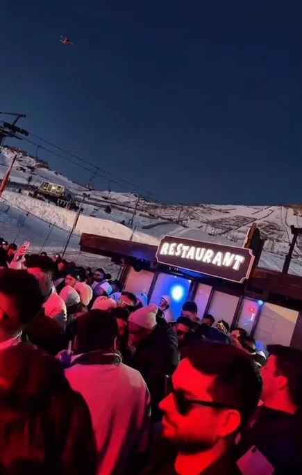 Pessoas em uma fila fora de um restaurante, com neve ao fundo, ao entardecer, com um avião no céu.