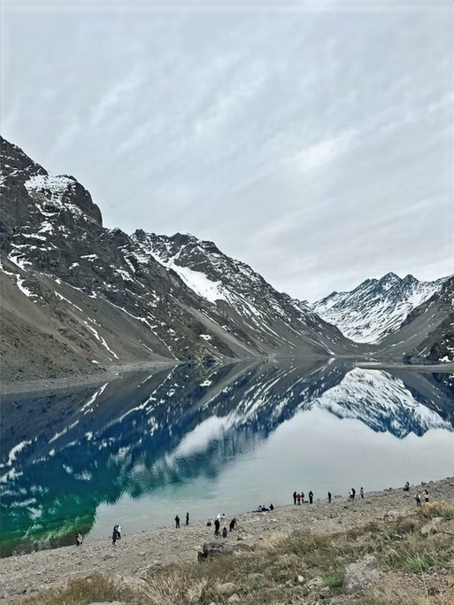 Lago de montanha com águas tranquilas e reflexo das montanhas ao redor, céu nublado e pessoas na margem.