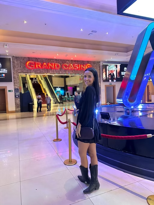 Mulher jovem sorridente em frente ao cassino Grand Casino, no interior de um shopping, com escadas rolantes ao fundo.