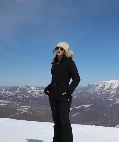 Mulher de óculos e gorro branco, vestindo casaco preto, posando na neve com montanhas ao fundo.