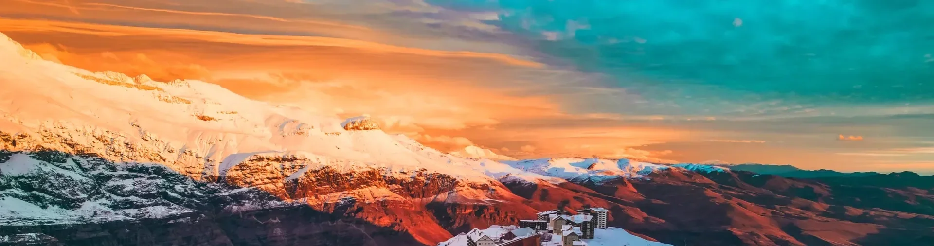 Montanhas com neve sob um céu colorido de pôr do sol, com prédios na parte inferior da cena.