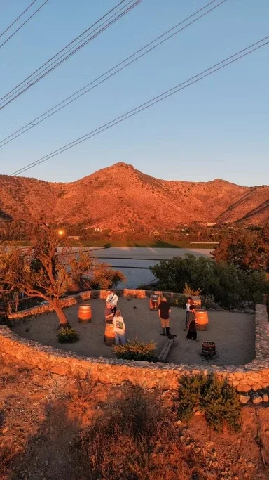 Grupo de pessoas com malas na plataforma de embarque de um aeroporto ao pôr do sol, próximo a um pequeno lago e montanhas ao fundo.
