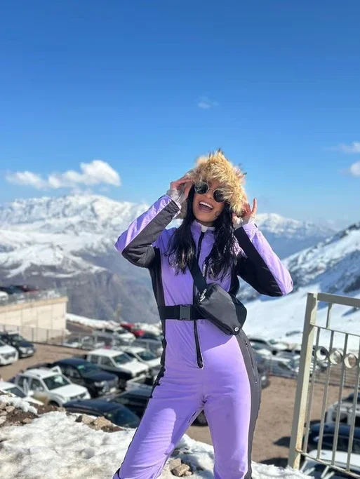 Mulher com roupa de esqui roxa, usando óculos de sol e chapéu com pelúcia, sorrindo em cenário de neve nas montanhas.