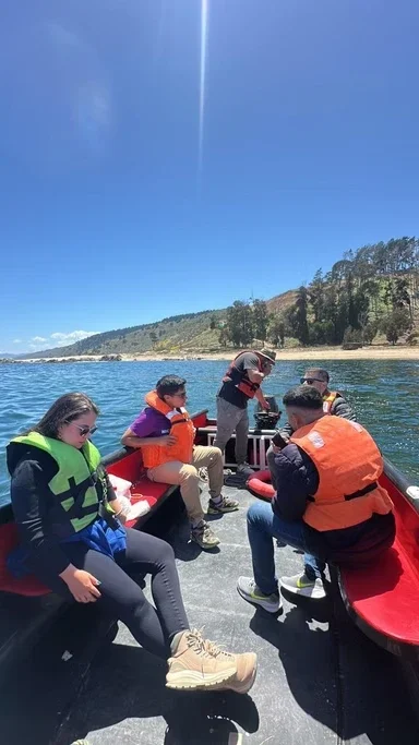 Pessoas em um barco no lago com o céu claro ao fundo, alguns usando coletes salva-vidas, uma mulher ajustando algo na frente e o cenário natural ao redor.