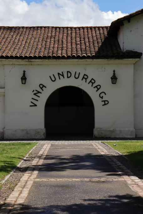 Entrada de um edifício com arco e sinais de iluminação nas paredes, com a inscrição "Vina Undurraga" acima da entrada, cercado por um caminho de pedra e grama.