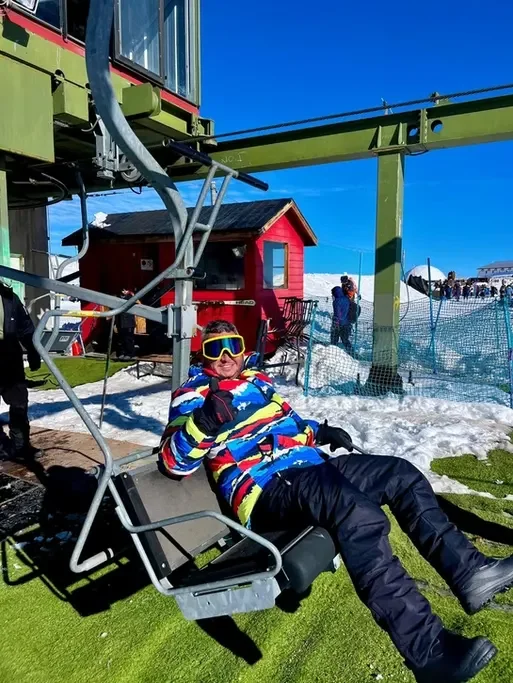 Pessoa de óculos de proteção e roupa de esqui relaxando em uma cadeira de teleférico na neve, com pista de esqui, redes de proteção e uma cabana vermelha ao fundo, em ambiente de estação de esqui.