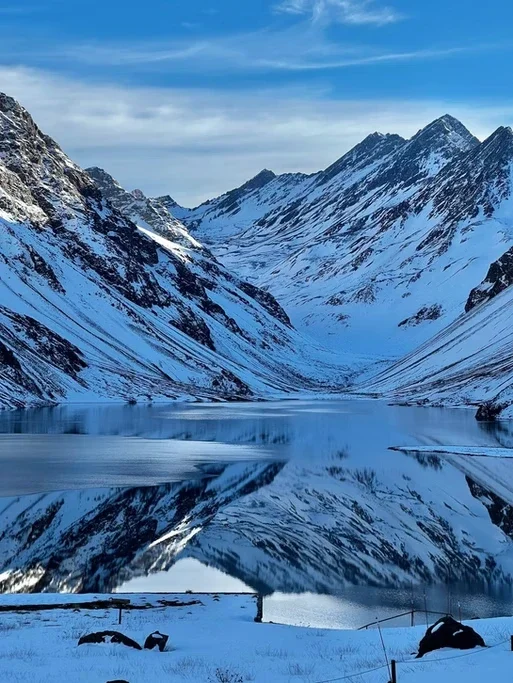Montanhas nevadas ao redor de um lago tranquilo com reflexo das montanhas na água.