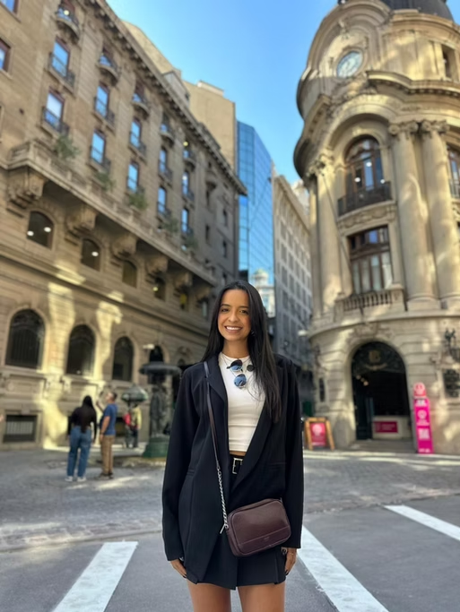 Mulher sorrindo em uma rua urbana com prédios históricos de arquitetura clássica ao fundo.