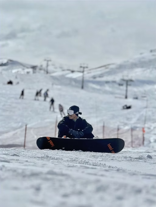 Pessoa com óculos de proteção e casaco escuro sentado na neve ao lado de um snowboard, em uma estação de esqui com outras pessoas esquiando ao fundo.