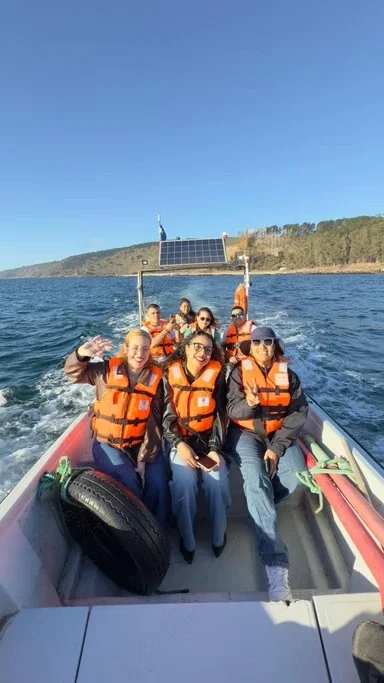 Grupo de pessoas em um barco no lago, usando coletes salva-vidas, com água, céu claro e uma paisagem de colinas ao fundo.