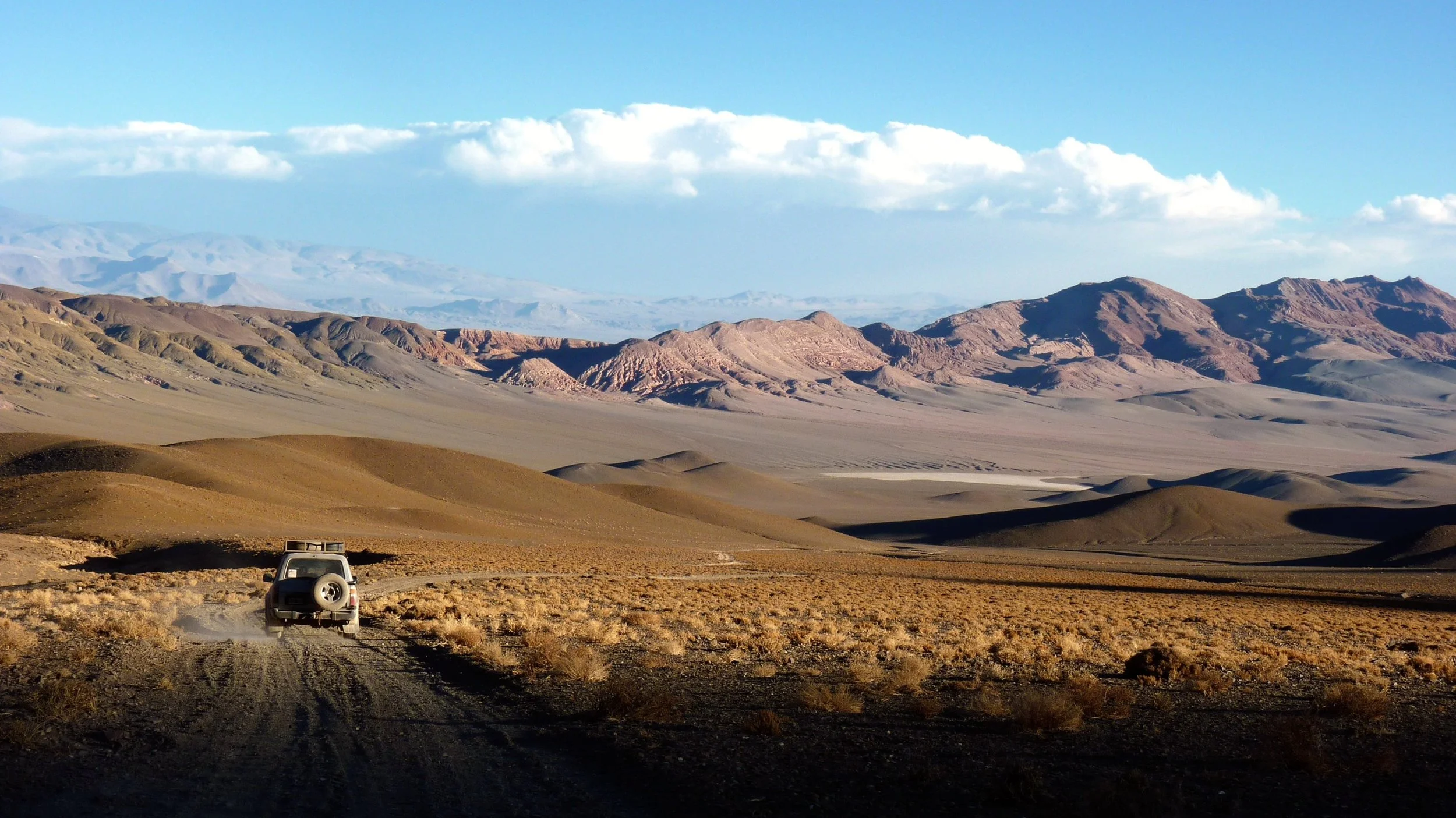 Paisagem desértica com montanhas ao fundo e carro dirigindo por uma estrada de terra.