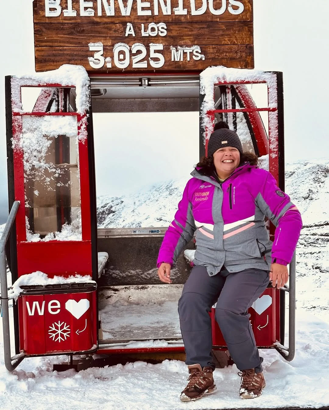 Pessoa sorridente sentada em uma cabine de teleférico de neve, vestindo roupas de inverno roxas e cinza, com fundo de montanhas nevadas.