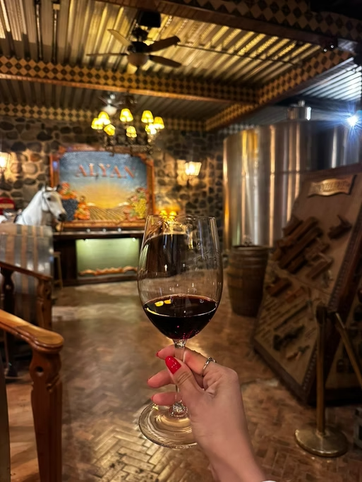 Vaso de vinho tinto sendo segurado por uma pessoa com unhas vermelhas em um ambiente rústico e aconchegante, decorado com pedra e madeira, com uma pintura de fundo, uma máquina de cerveja e uma área de jantar.