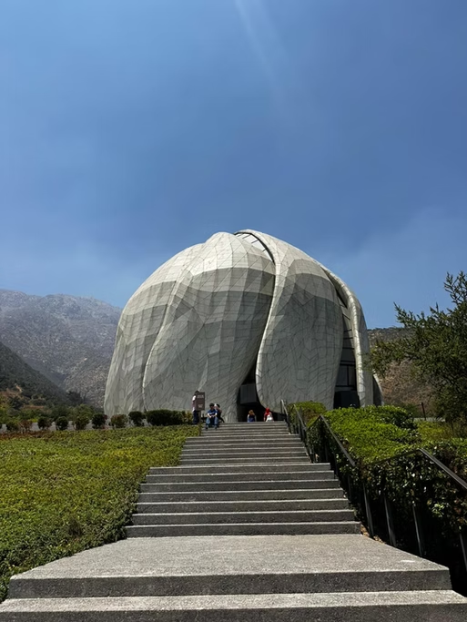 Edifício futurista de forma ovular com uma escadaria de acesso, rodeado por natureza e montanhas ao fundo, sob céu claro.