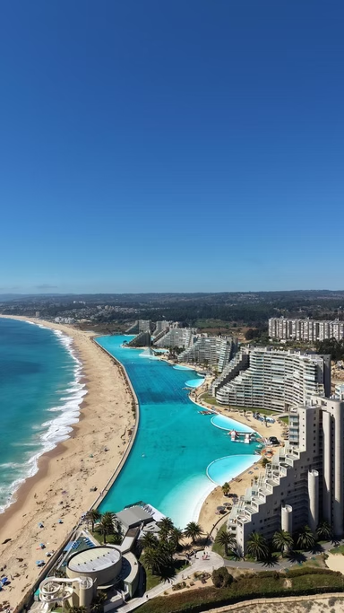 Vista aérea de uma praia com mar azul, um Hotel de luxo com piscinas de borda infinita, e prédios na orla, sob céu claro e ensolarado.