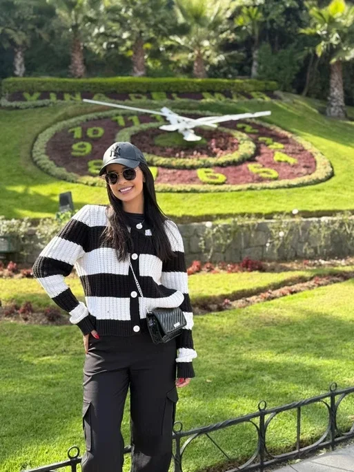 Mulher sorridente com óculos e boné, vestindo suéter listrado preto e branco, posando em frente a um relógio floral ao ar livre.