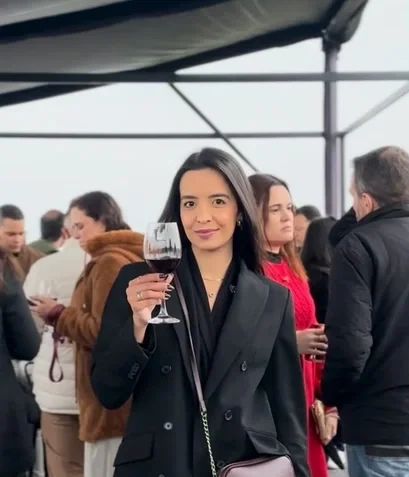 Mulher sorridente segurando taça de vinho em evento ao ar livre com várias pessoas ao fundo