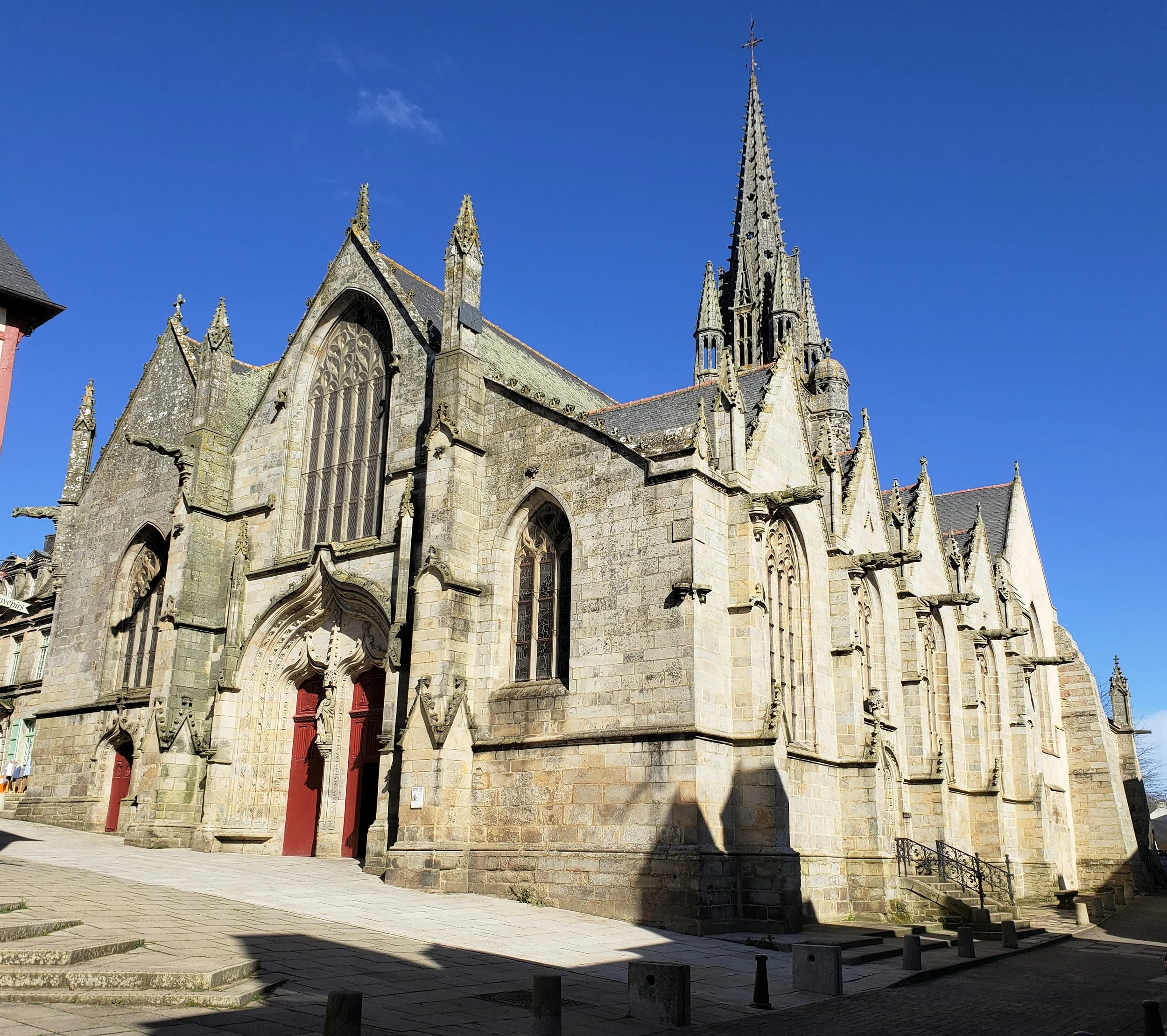 The magnificent gothic Basilique Notre-Dame du Roncier in the town center of Josselin, France, its ornate stone facade, flying buttresses and soaring spire set against a brilliant blue Breton sky.