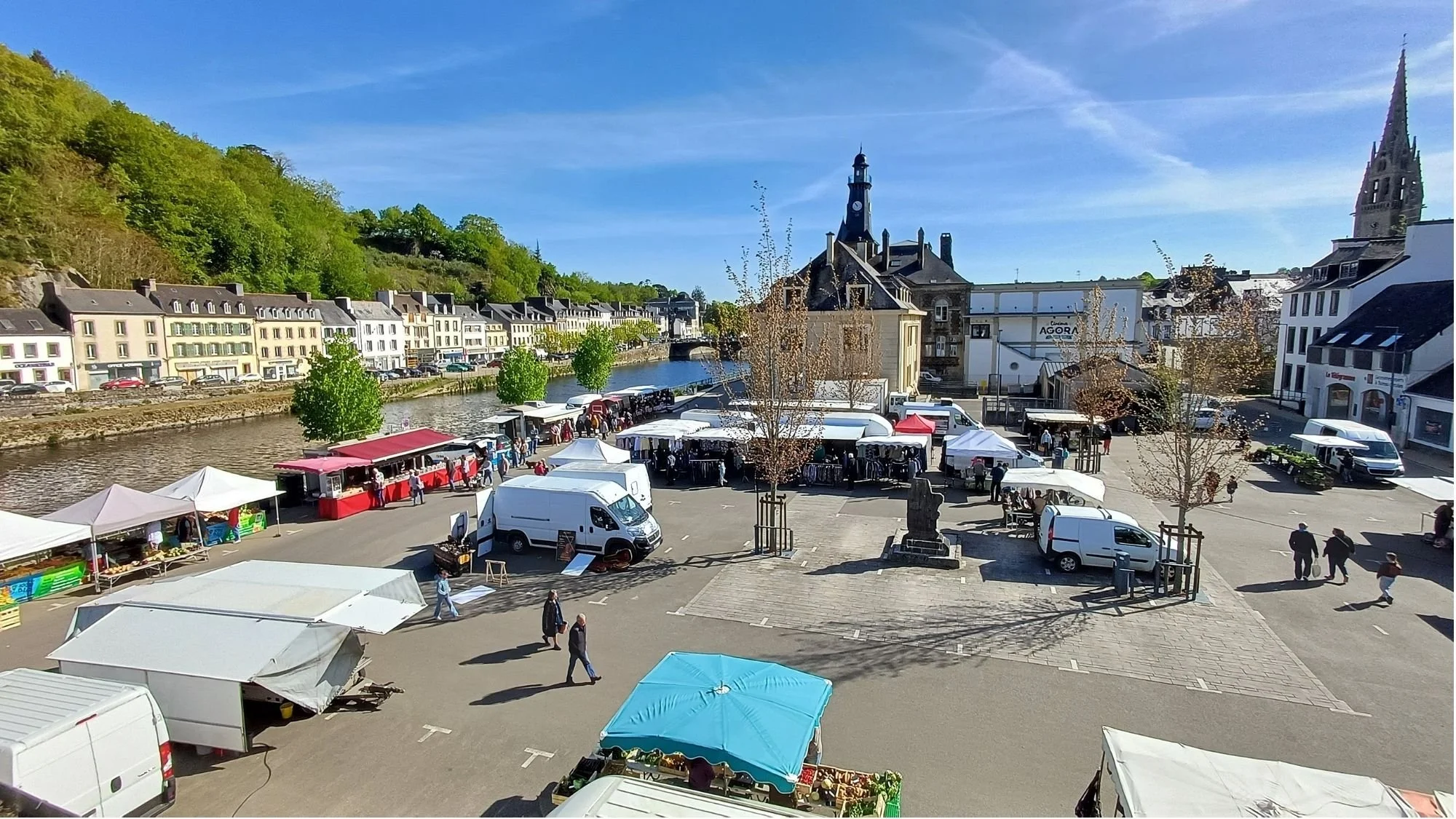 View of the Châteaulin, France weekly market from the Numéro 1 B&B with the Aulne River and the steeple of Église Saint-Idunet visible under a blue sky.