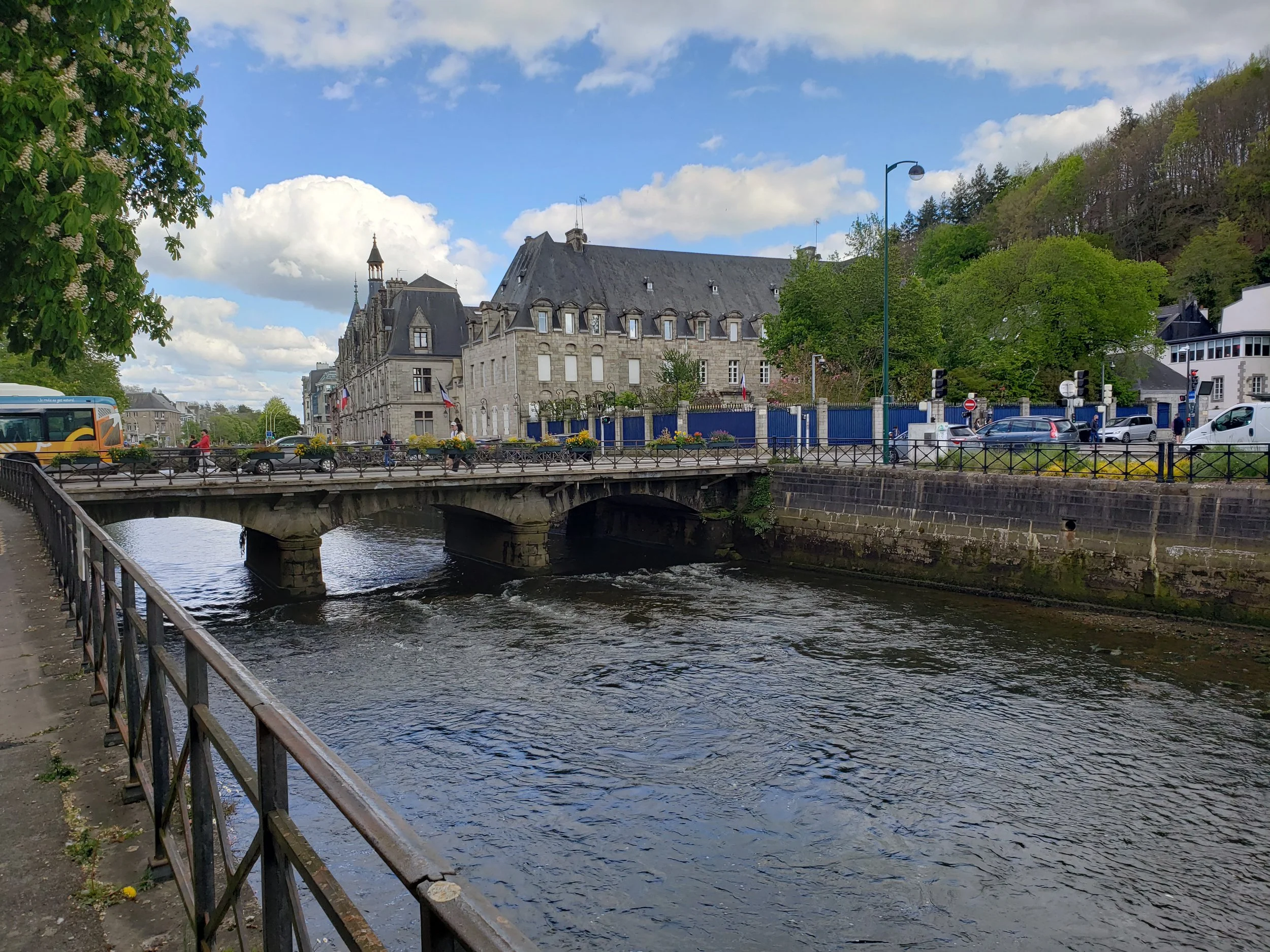 A flower-lined footbridge over the Odet River in Quimper, France, with old stone buildings and the trees of Mont Frugy in the background.