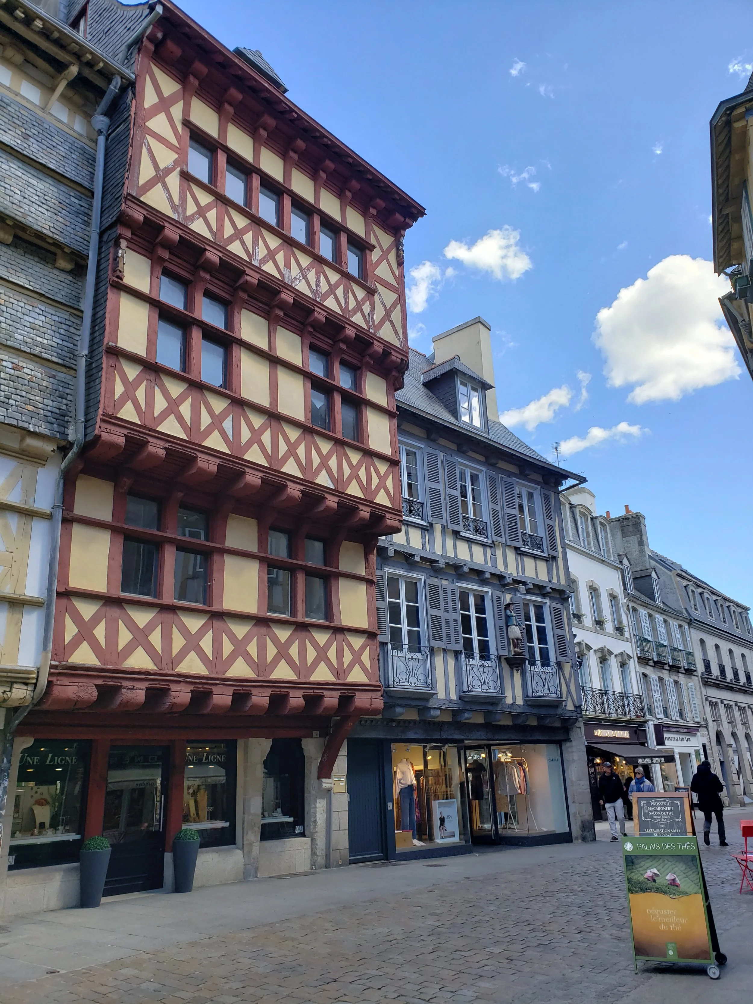 Tall half-timbered Tudor-style buildings lining the cobblestone Rue Kéréon in the old town of Quimper, France, housing bistros, crêperies and boutiques at street level beneath their historic upper floors.