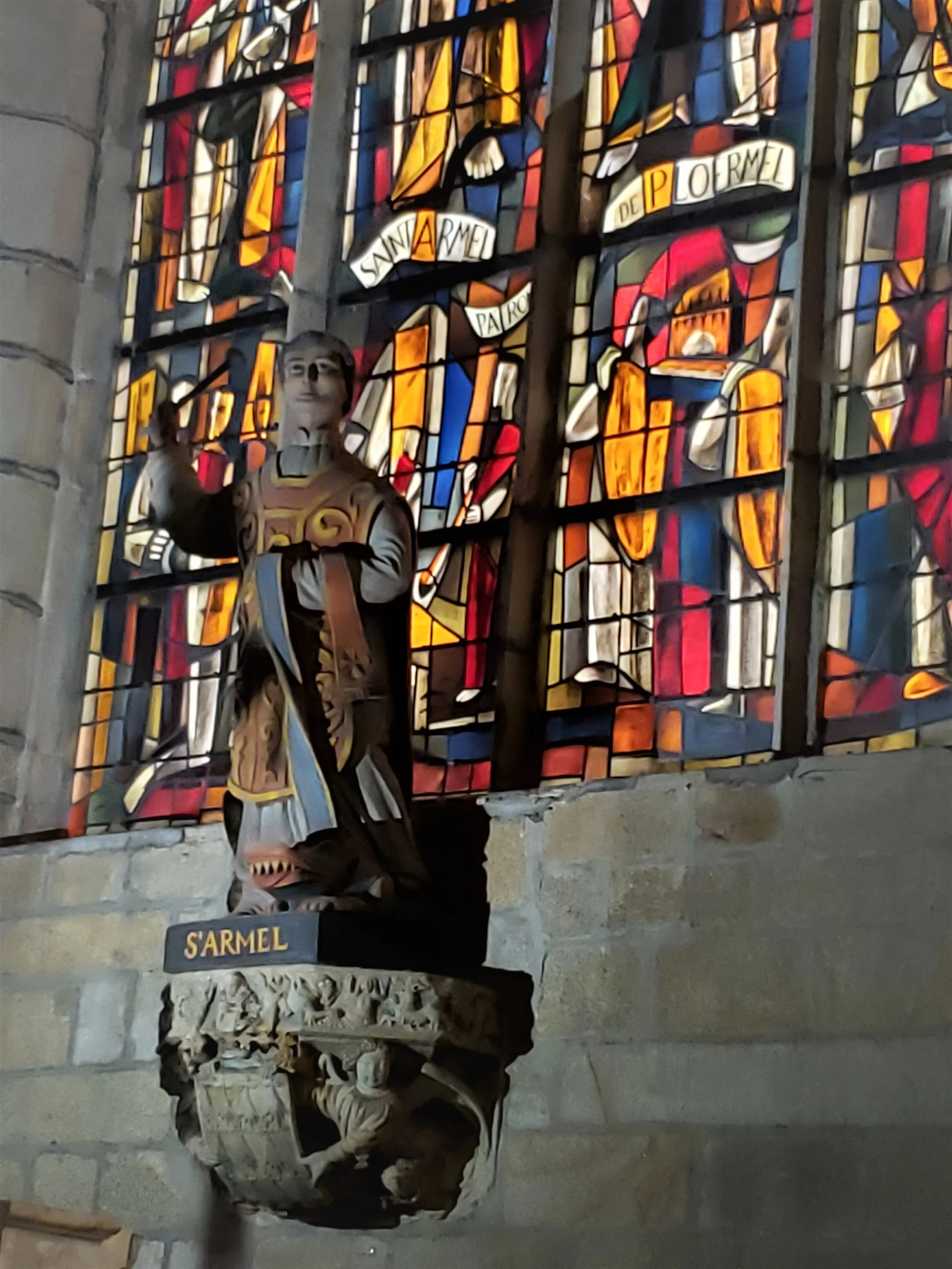 A sculpted statue of Saint Armel standing proudly before a stained glass window bearing his name inside the historic Église Saint-Armel church in Ploërmel, France.