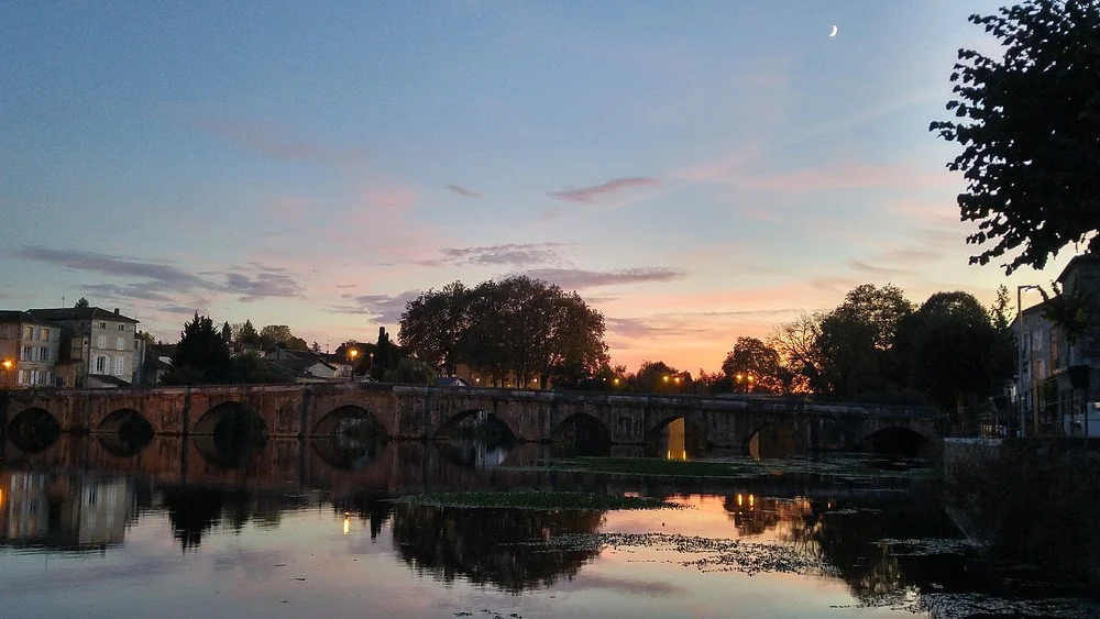 An early evening view of the Pont Vieux, the 14th century old bridge spanning the Vienne River in Confolens, France, seen from the peaceful west bank.