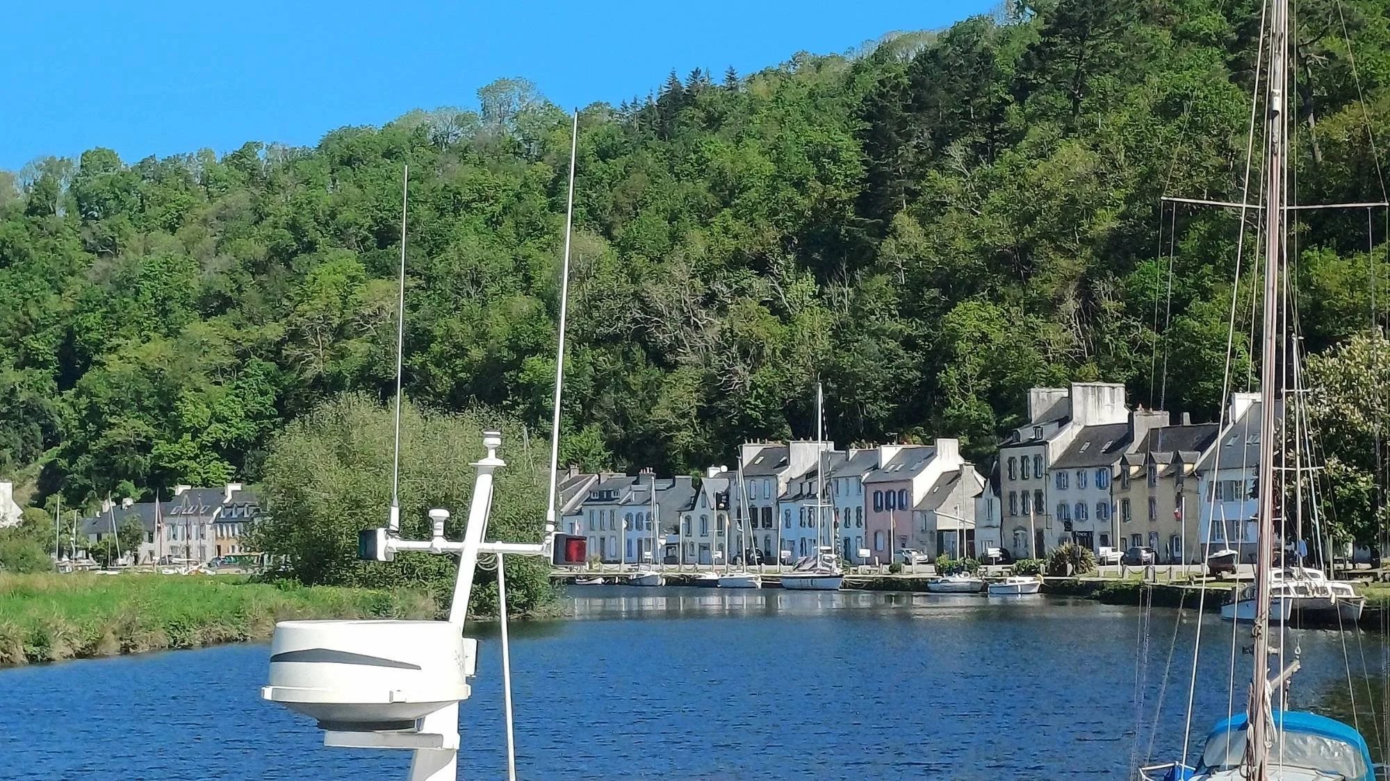 Boats moored along Quai Amiral Douguet on the Aulne River in Port-Launay, France, with riverside buildings and tree-covered hills under a bright blue sky.
