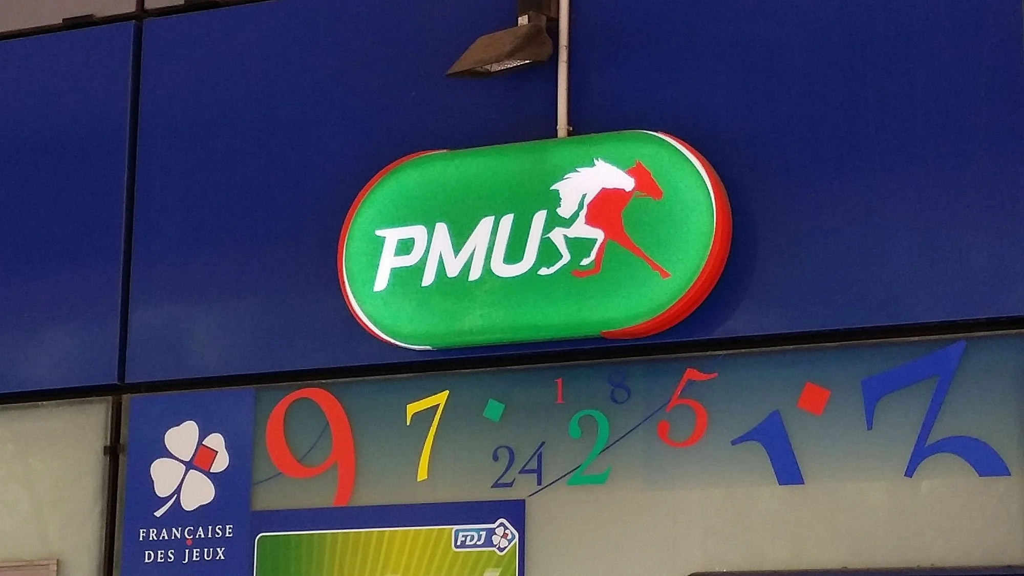 Exterior wall-mounted PMU horse racing sign with a Française des Jeux (FDJ) sign below on a building facade in France.
