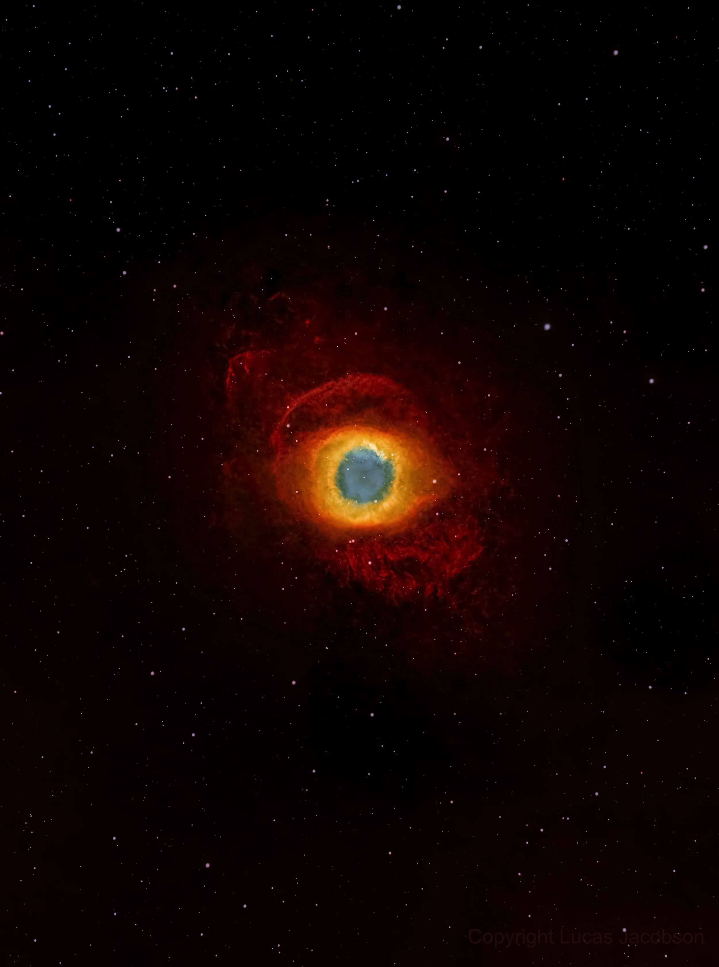 A colorful nebula with a bright, glowing ring at its center, surrounded by red and orange gaseous clouds, set against a backdrop of numerous small stars in the night sky.