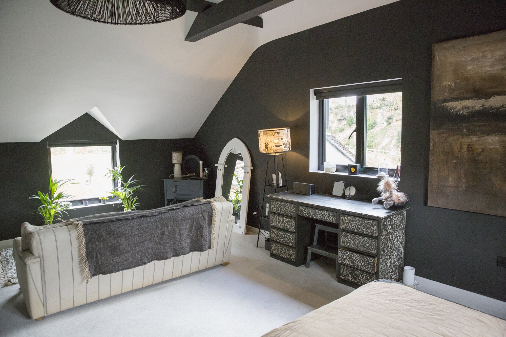 An attic bedroom with dark walls and sloped ceiling, featuring a white bed with a gray blanket, two plants near a window, a vintage-style dresser with decorative drawers, a lamp, and a teddy bear on the dresser. There is a large framed artwork on the