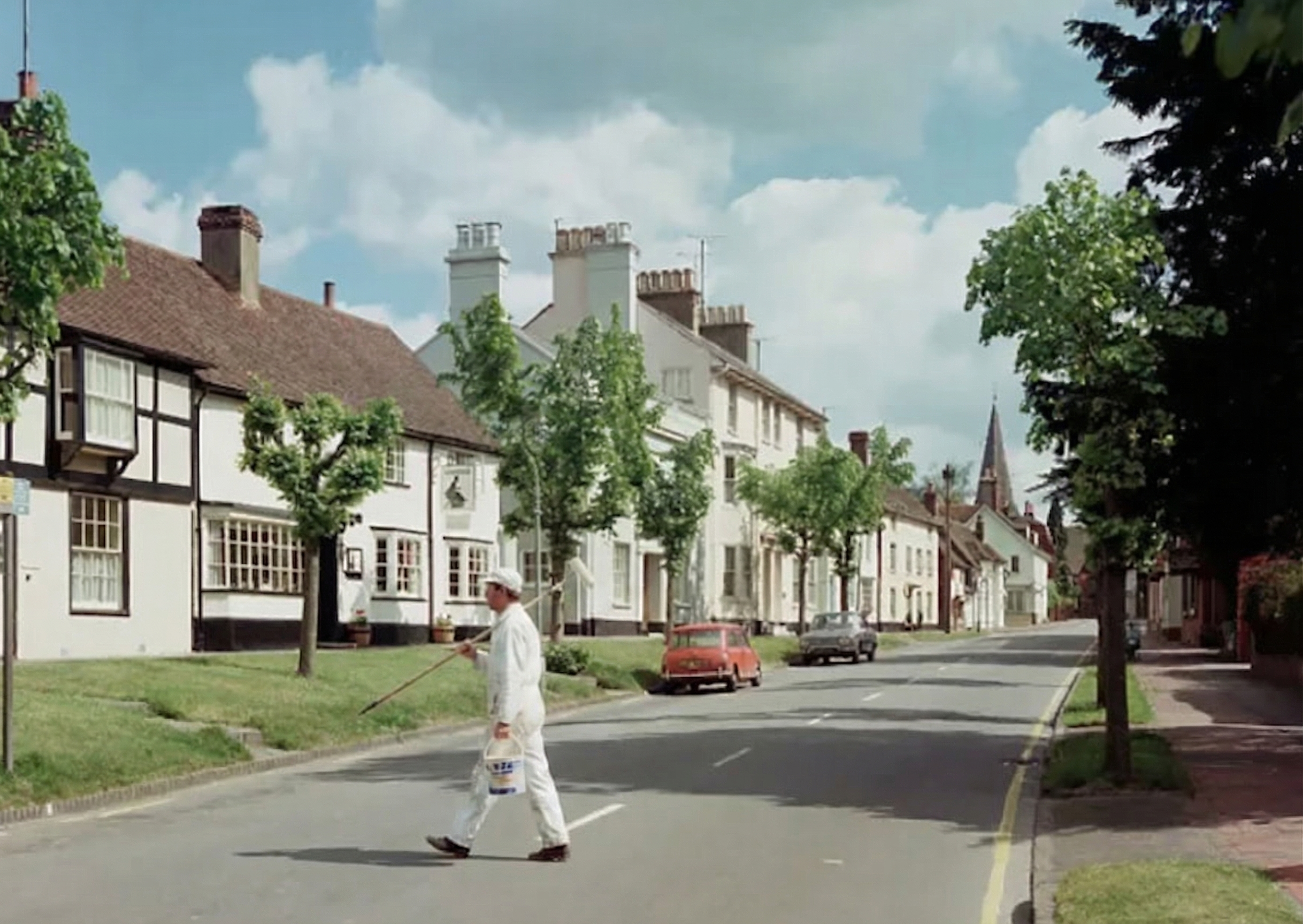 A man in white clothing painting the curb on a residential street using a paint roller, with cars parked alongside and houses with chimneys and trees lining the street under a partly cloudy sky.