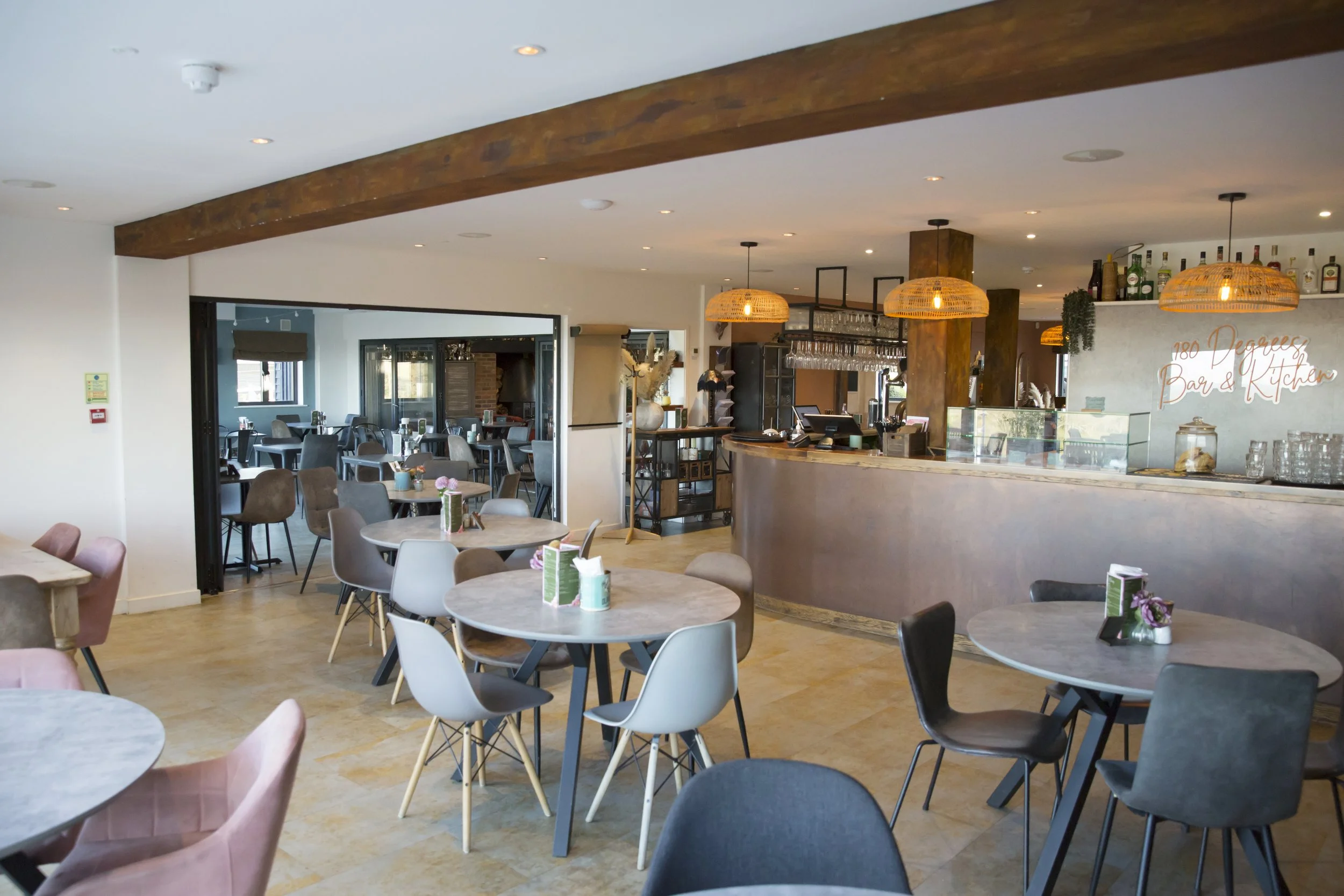 Interior of a modern restaurant or cafe with round tables and assorted chairs, a bar area with hanging glasses, bottles for alcohol, and warm lighting fixtures.