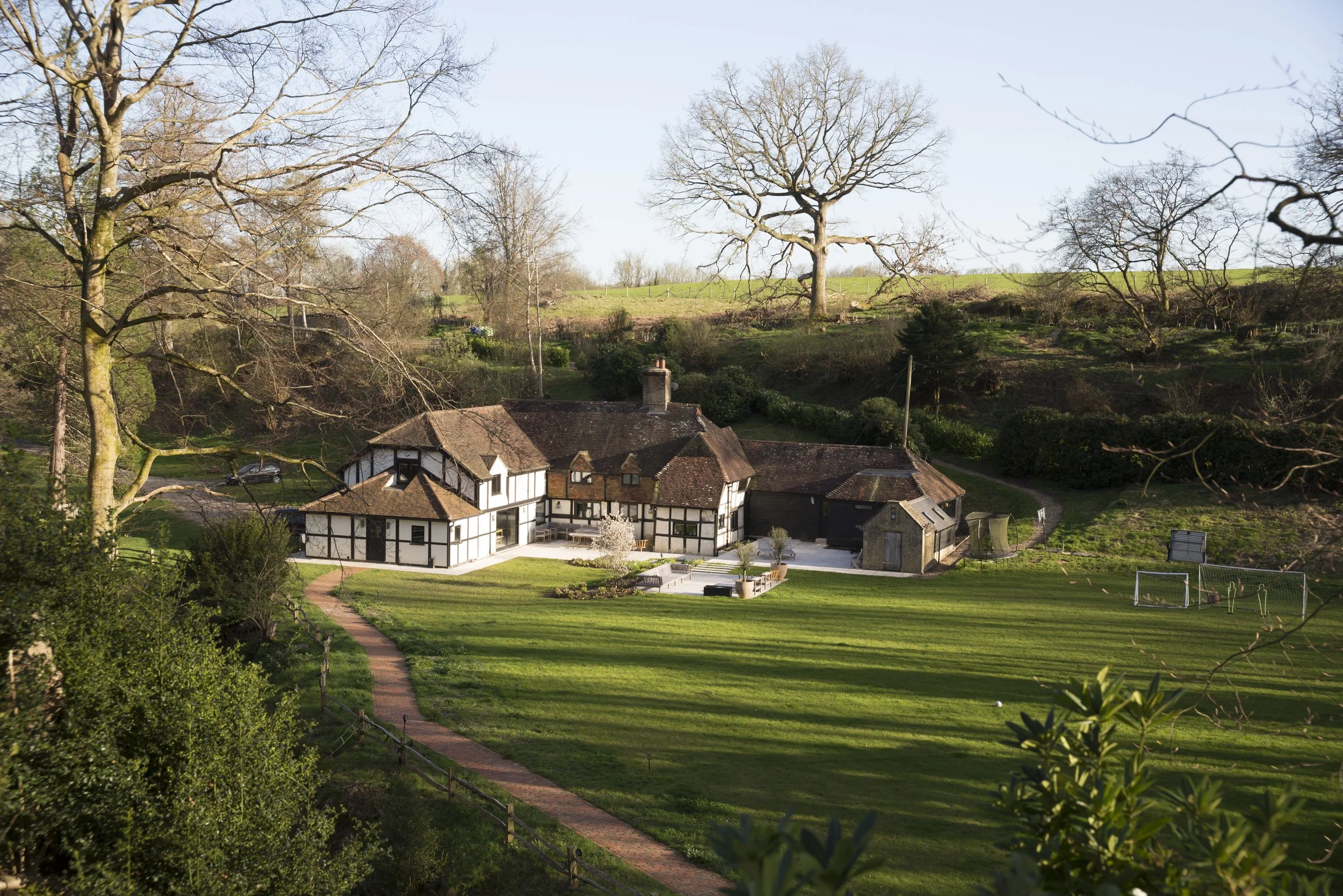 A house surrounded by green lawn and trees, with a pathway leading to the house. The house has a Tudor-style design with black and white exterior and a brick chimney. In the background, there are more trees and a hill with sparse vegetation. The sky 