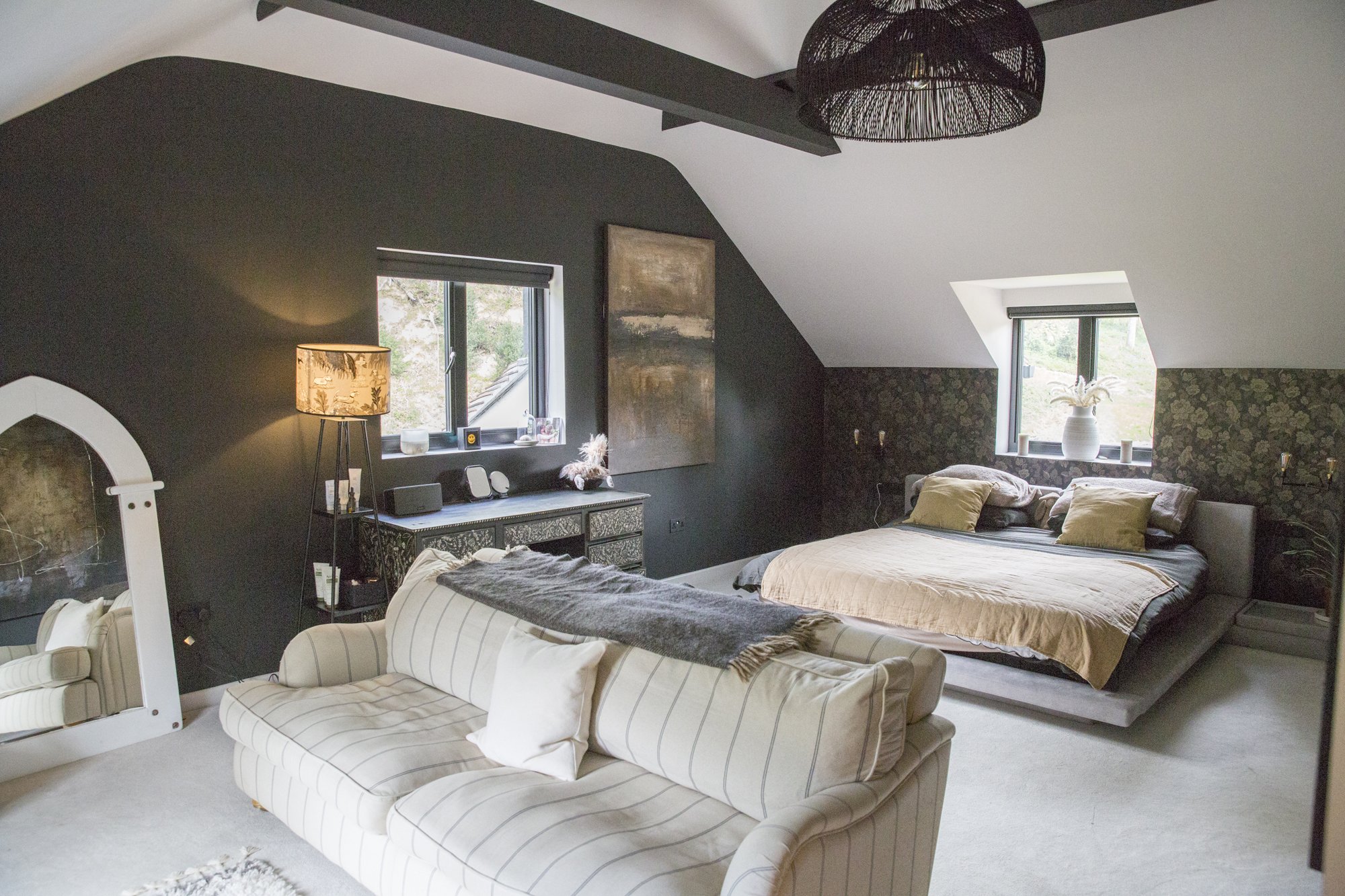 A bedroom with sloped ceilings and two windows, decorated in a modern style with dark and neutral colors. There are two beds, a beige chair, a mirror, a black side table, and various decorative items like a lamp, artwork, and vases.