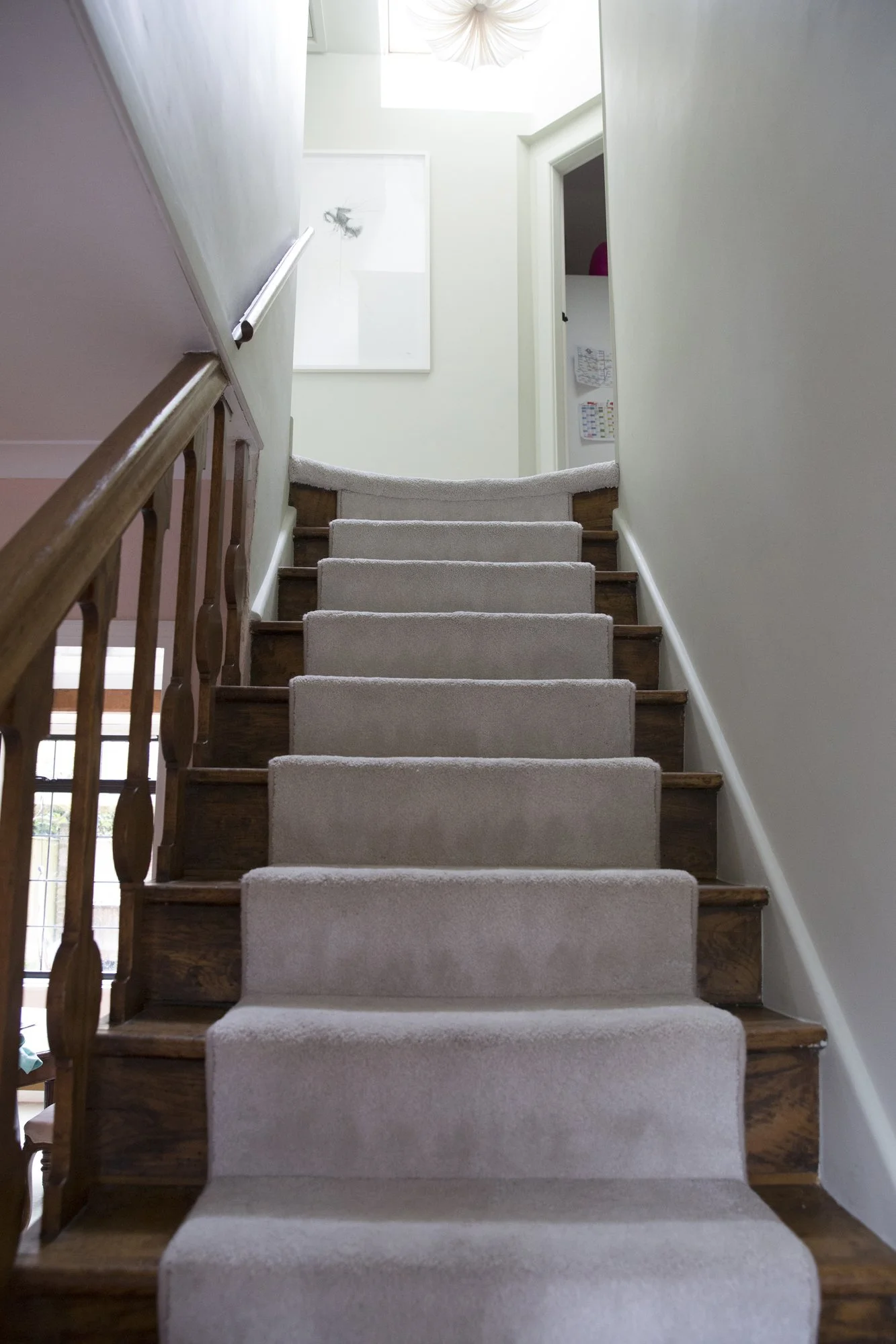 Interior view of a staircase with beige carpeted steps, wooden handrails, and white walls, leading up to a bright area with a white framed artwork and an open door on the right side.